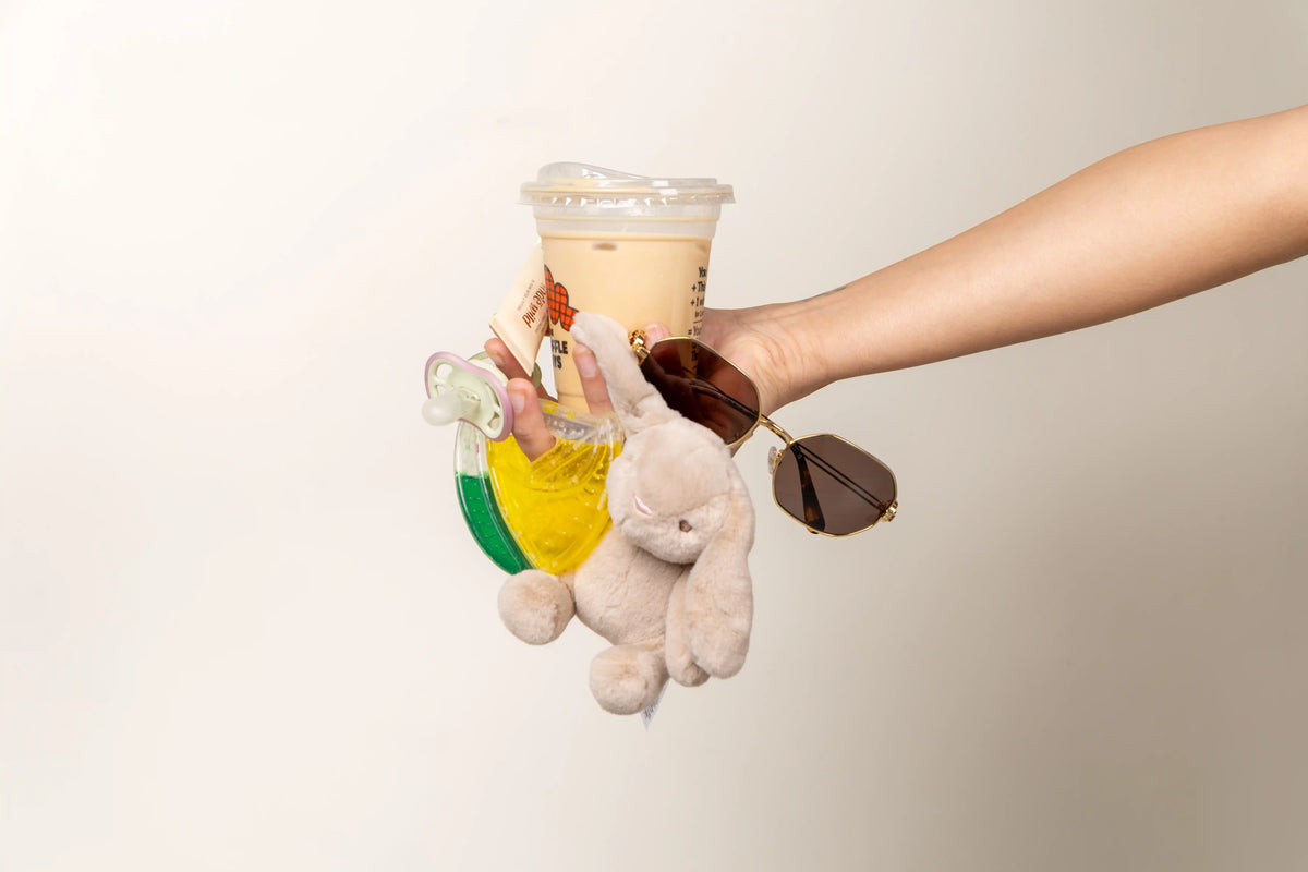 A hand holding a clear plastic takeaway coffee cup with a lid, attached to a keychain featuring a small beige plush bunny, brown sunglasses, a yellow and green bottle, and a pacifier, against a plain light beige background.
