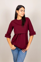 A woman modeling a three-quarter sleeve peplum top in deep burgundy, paired with blue jeans, against a plain light gray studio background.