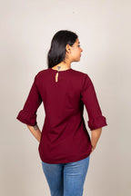 A woman viewed from the back wearing a maroon three-quarter sleeve top with rolled cuffs and a small keyhole cutout at the neckline, paired with blue jeans, against a plain light beige background.