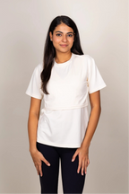 A woman modeling a white short-sleeve top with a flap on the front to make breast feeding easier, paired with black pants, against a plain light gray studio background.