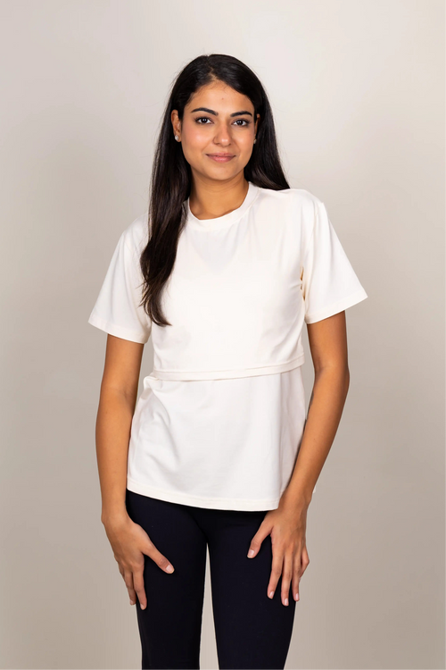A woman modeling a white short-sleeve top with a flap on the front to make breast feeding easier, paired with black pants, against a plain light gray studio background.