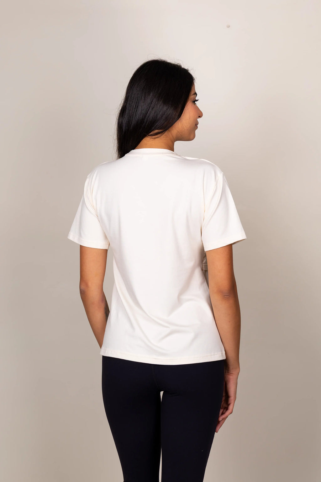 A woman viewed from the back wearing a plain white short-sleeve crewneck t-shirt and dark navy leggings, standing against a neutral beige studio background.